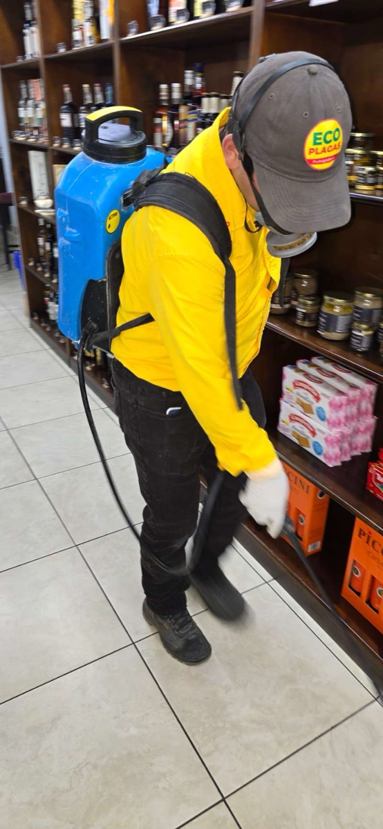 Técnico de EcoPlagas uniformado con gorra, respirador y bomba rotulada durante un servicio comercial